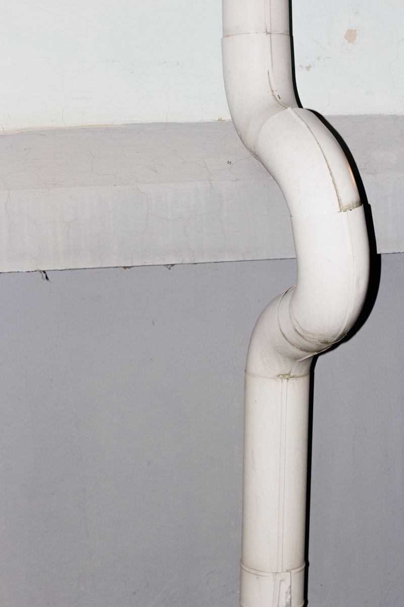 White PVC vent pipe with an offset bend mounted on a concrete exterior wall, similar to the exterior run of a radon mitigation system